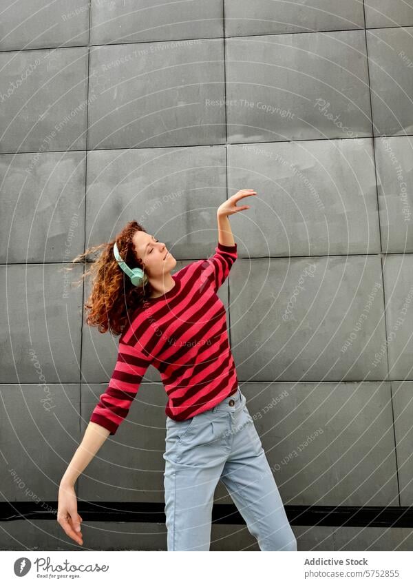 Young redhead woman in striped shirt enjoys music outdoors young headphone dance urban wall gray carefree rhythm favorite song enjoyment leisure casual