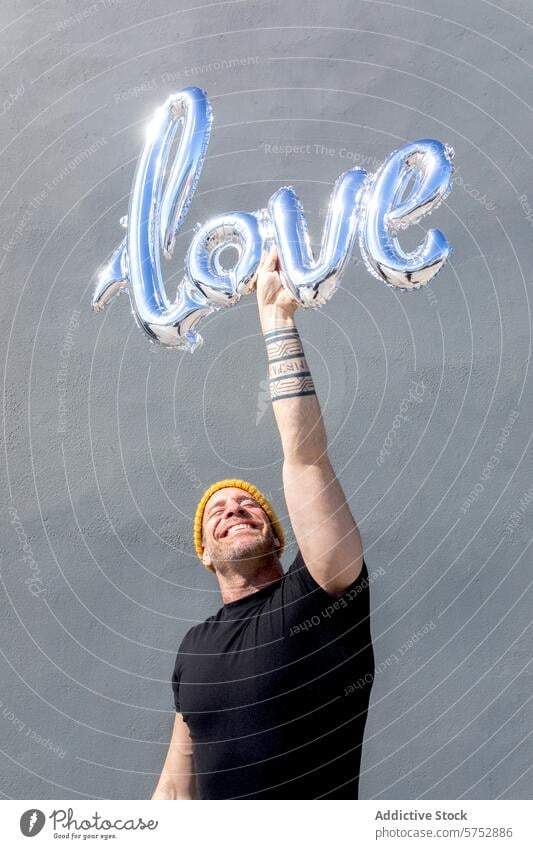 Joyful man holding love balloon letters joy happy beanie yellow gray background celebration affection shiny metallic spell emotion positive smile cheerful bliss