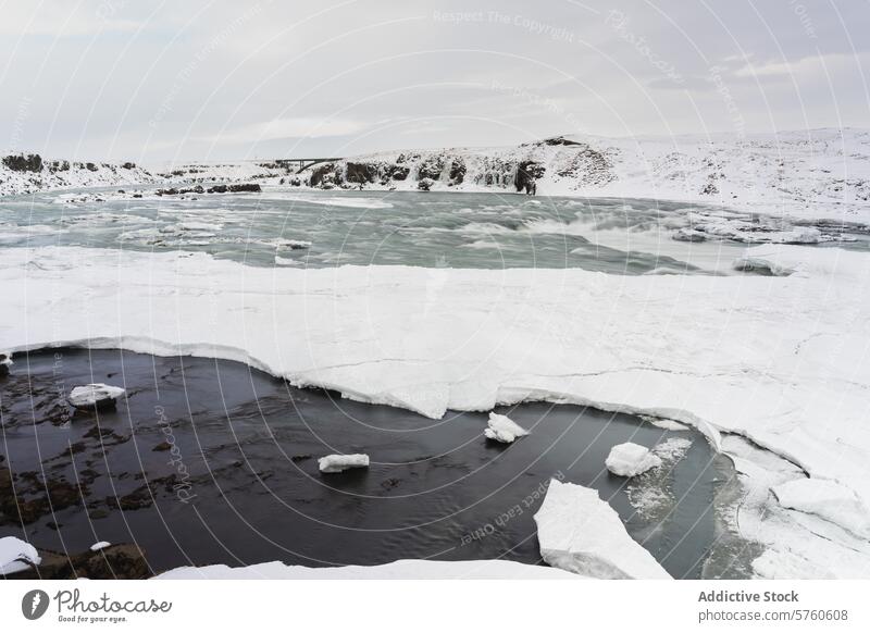 The stark beauty of a partially frozen river winding through a snow-laden Icelandic landscape, under a overcast winter sky ice nature outdoor travel cold water