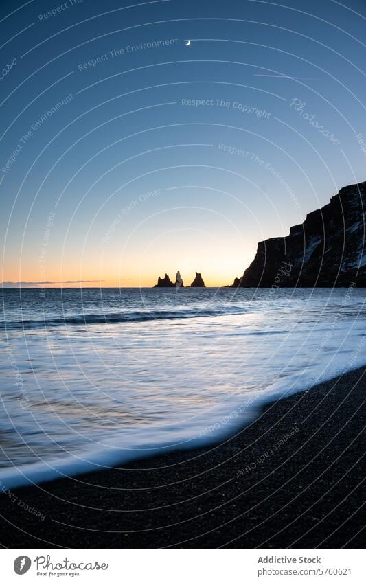 The crescent moon watches over Reynisdrangar seastacks, as twilight caresses the shores of Iceland's iconic black sand beach dusk ocean silhouette evening sky