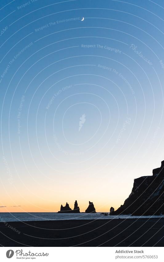 Under the crescent moon, the silhouettes of Reynisdrangar sea stacks stand tall against the twilight sky on Iceland’s black sand beach serenity cliffs dusk