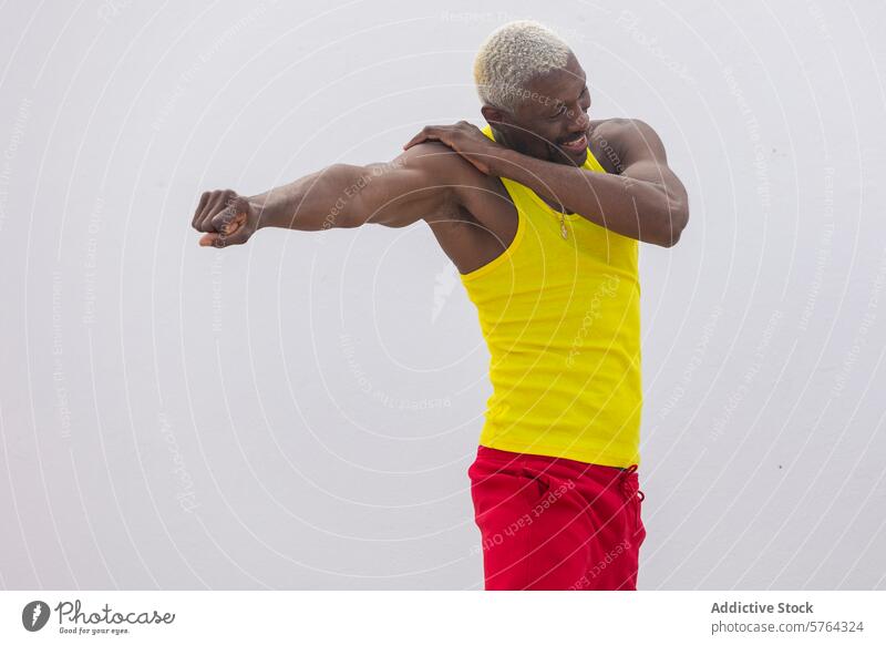 A cheerful man in a bright yellow tank top and red shorts stretches his arm, showcasing a healthy, active lifestyle fitness stretching exercise outdoors