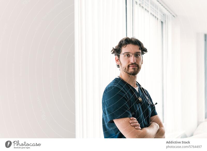 A serene male medical professional stands with folded arms in a bright, contemporary clinic setting, exuding calm competence healthcare scrubs stethoscope