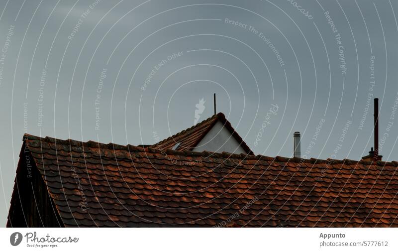 View of the roof surface and ridge of an old saddle roof covered with red plain tiles against a light gray sky. A roof peak with a so-called interception device of a lightning protection system (lightning rod)