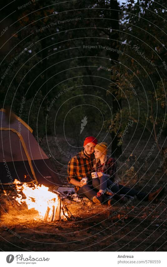 Young couple a guy and a girl in bright knitted hats stopped at a camping young beard warmth tent bonfire love get warm drink tea firewood blue orange cage