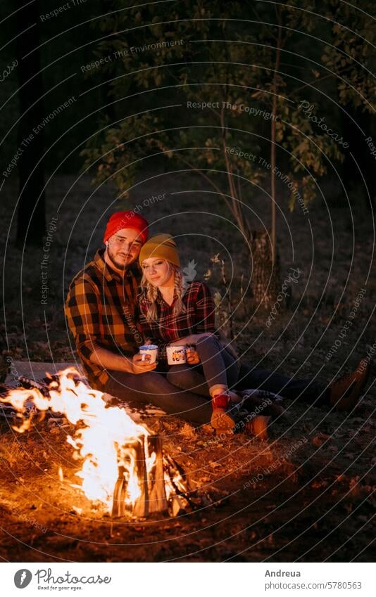 Young couple a guy and a girl in bright knitted hats stopped at a camping young beard warmth tent bonfire love get warm drink tea firewood blue orange cage