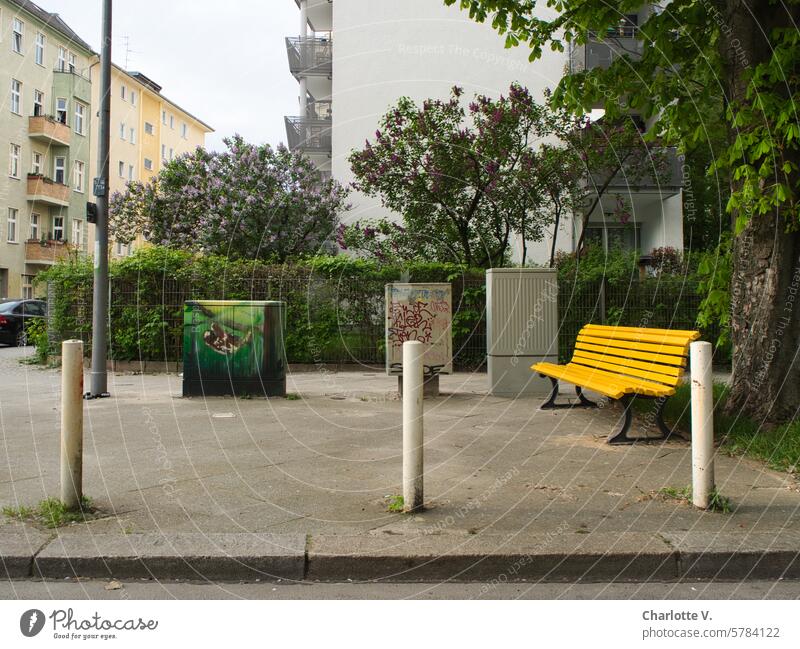 Platz mit wenig Aufenthaltsqualität I Stillleben mit Versorgungskästen, einer gelbe Bank und drei Pollern urbanes Stillleben absurd trist Stadtgrün