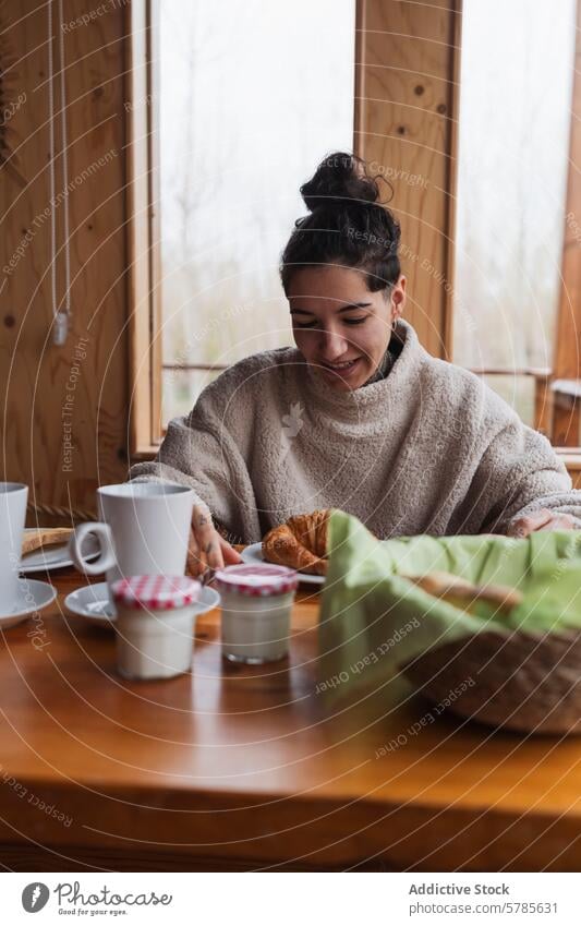 Woman relishing a cozy breakfast on a chilly morning woman smiling wooden table window enjoy scene happy croissant coffee yogurt bread jam warm sweater indoor