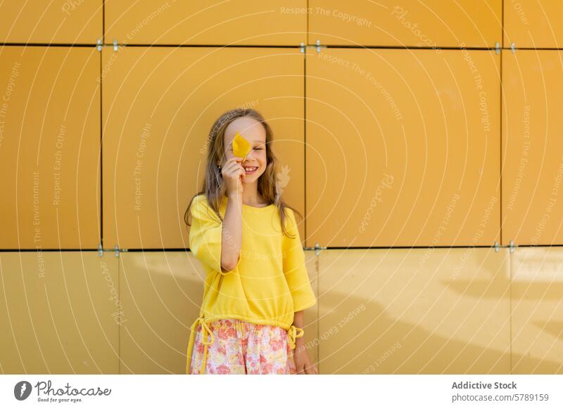 Young girl playing with a leaf in autumn colors child playful yellow happy smile wall bright standing outfit cute joy fun casual season cheerful nature