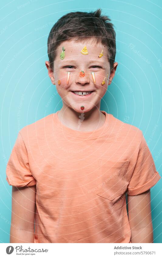 Cheerful boy with fruit stickers on face and teal background smile joyful blue background adornment colorful happy child youth playful freshness decorative