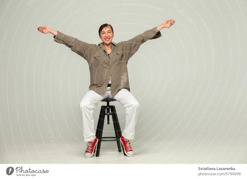 A middle-aged woman in casual clothes and red sneakers poses with a smile in the studio against a gray background with her arms outstretched. people one person