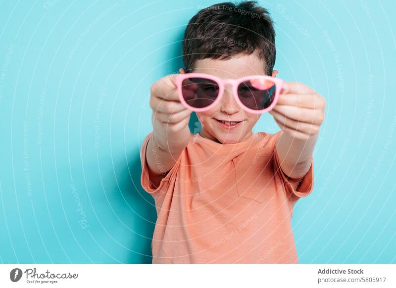 Smiling boy holding pink sunglasses against a blue backdrop child kid student school background fun playful cheerful happy joy casual clothing fashion accessory