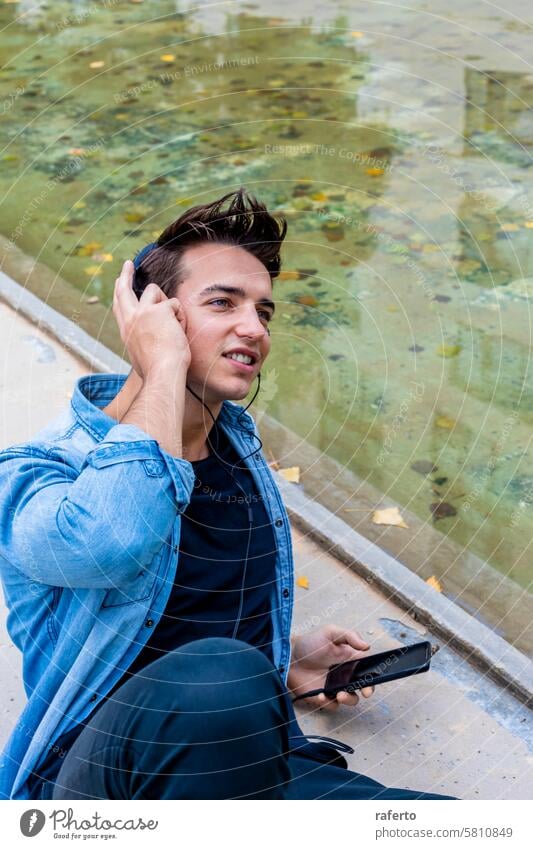 Man listening to music by a pond. man headphones smartphone denim shirt black pants young adult focused relaxed urban outdoor sitting casual technology mobile