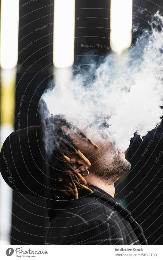 Black man exhaling cloud of smoke on street informal style dreadlocks urban tobacco contemporary cool nicotine fume habit city toxic town casual hat