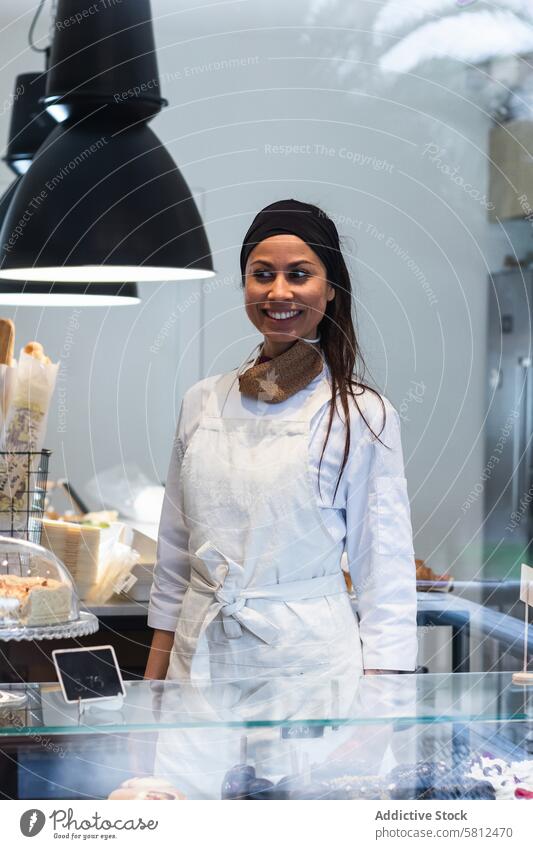 Cheerful woman standing at counter in bakery bakehouse showcase smile hospitality stall staff female work uniform professional job dessert confectionery vegan