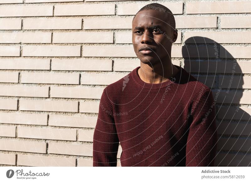 Young African American male standing near billet wall man casual serious style masculine ponder shadow brick wall town alone black knitted garment denim