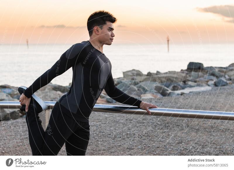 Ethnic athlete doing stretching exercise on seashore man workout fitness training sporty warm up sunset asian ethnic male sportsman leg lifestyle wellness young