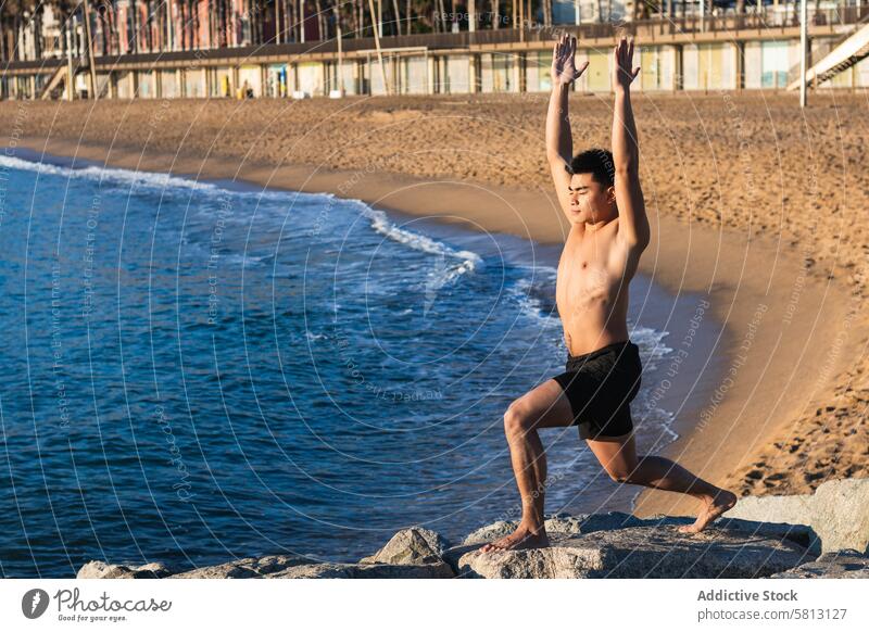 Ethnic man doing yoga pose on seashore beach warrior pose virabhadrasana practice balance stand male asian ethnic shirtless young lifestyle arms raised wellness