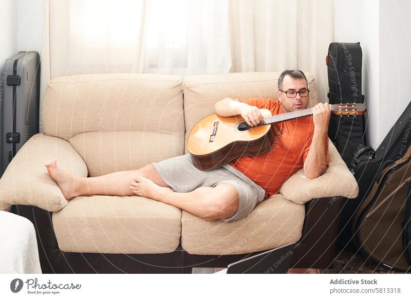 a man relaxing on the couch playing guitar laptop online lessons learning harmonics fun songs musician musical sitting adult computer internet male home