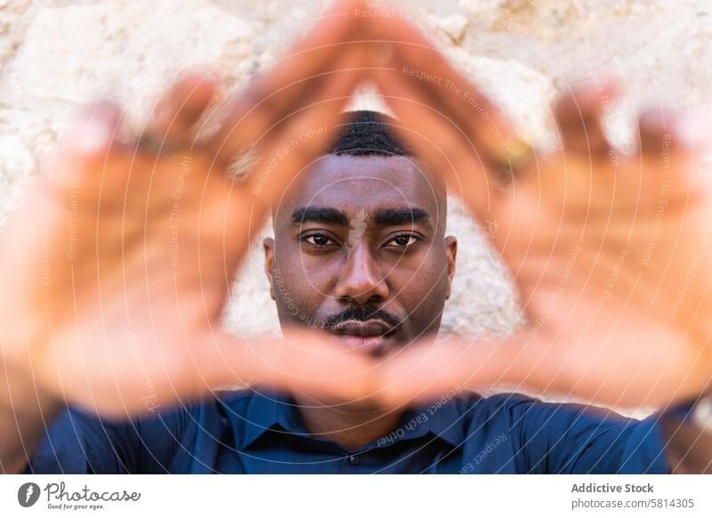 Black man showing triangle gesture with hands near wall serious demonstrate hands folded street confident alone african american black male adult shirt fence