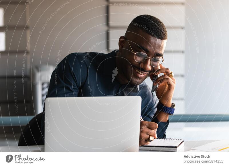 Black man talking on phone near laptop in office phone call take note notepad smartphone smile table document african american black male positive workplace