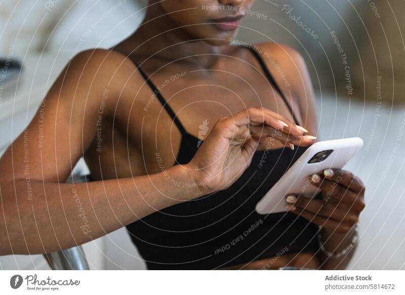 Anonymous black woman using smartphone while sitting on chair internet online text message social media watch scroll connection female device surfing mobile