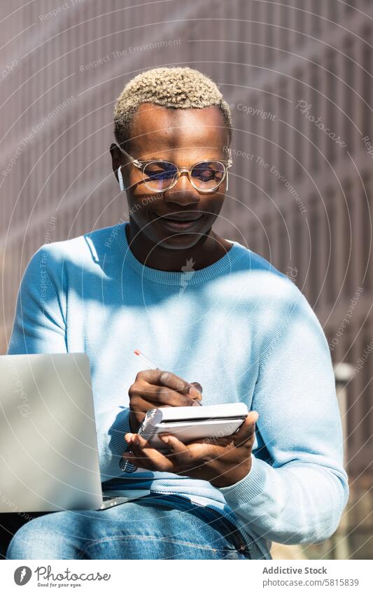 Positive young black male journalist taking notes during distance work on netbook man take note laptop planner freelance remote positive trendy city street