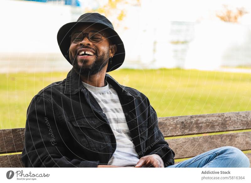 Trendy black man sitting on bench hipster happy park cheerful trendy hat adult african american ethnic male smile modern online internet communicate lifestyle