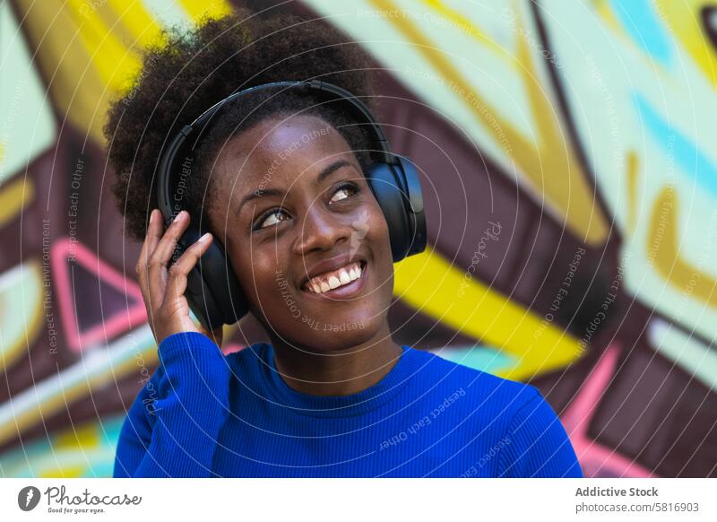 Positive ethnic woman enjoying music in city headphones listen graffiti wall cool millennial urban female black african american afro hairstyle cheerful happy