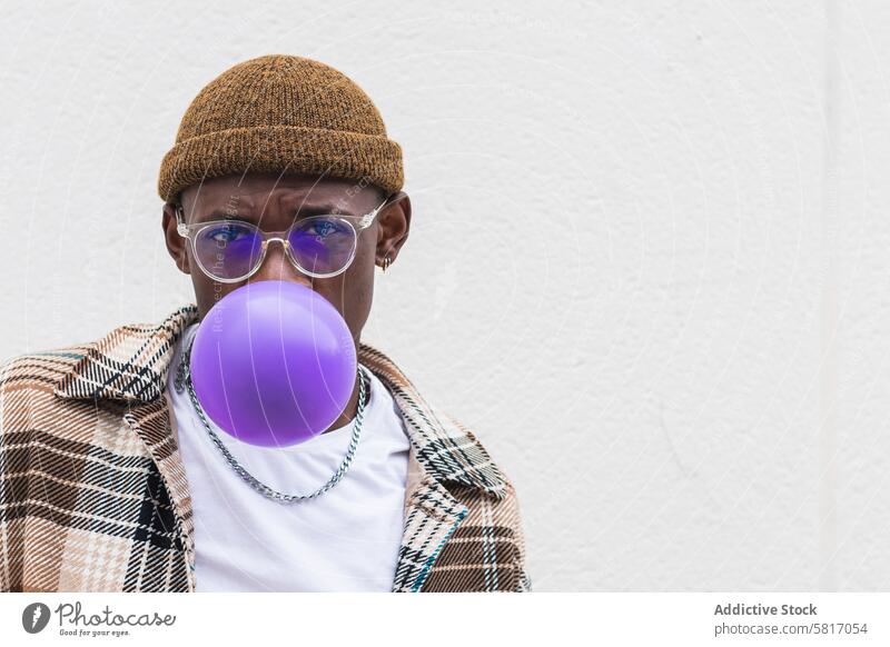 Trendy ethnic man blowing balloon style urban street casual young city modern male black african american hat glasses fashion outfit vivid hipster vibrant wall