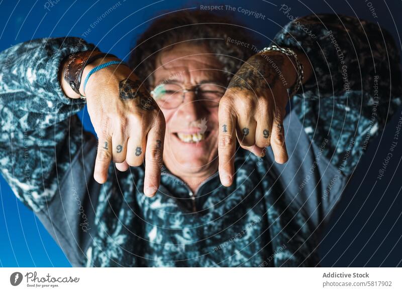 Positive tattooed mature man showing rock and roll gesture in studio cheerful smile happy informal positive joy glad rebel male middle age trendy curly hair