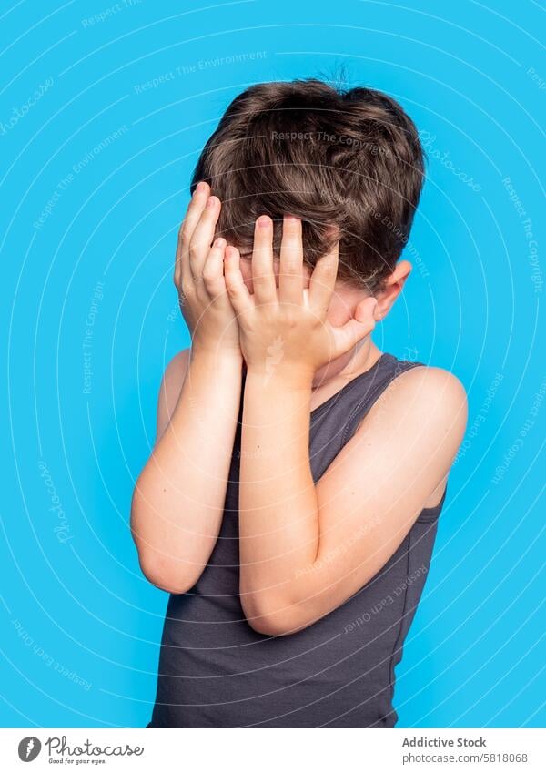 Young boy hiding face with hands on blue background child studio shot young looking away distressed upset playing emotion expression cover embarrassment shy