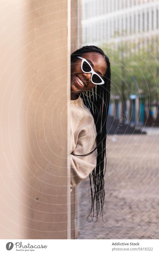 Cheerful ethnic woman peeking from behind column street smile city style fun building young female happy black african american cheerful trendy urban outfit