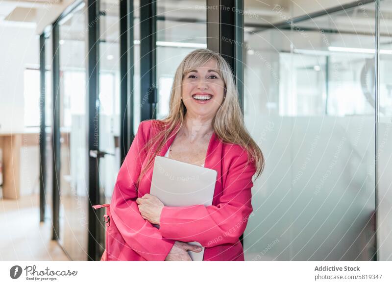 Confident businesswoman with laptop in modern coworking space professional smiling confidence success office stylish glass wall pink blazer holding standing