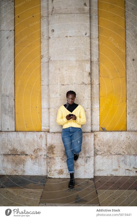 Young black woman texting on smartphone in urban setting concrete wall yellow sweater denim jeans standing absorbed technology mobile device casual fashion