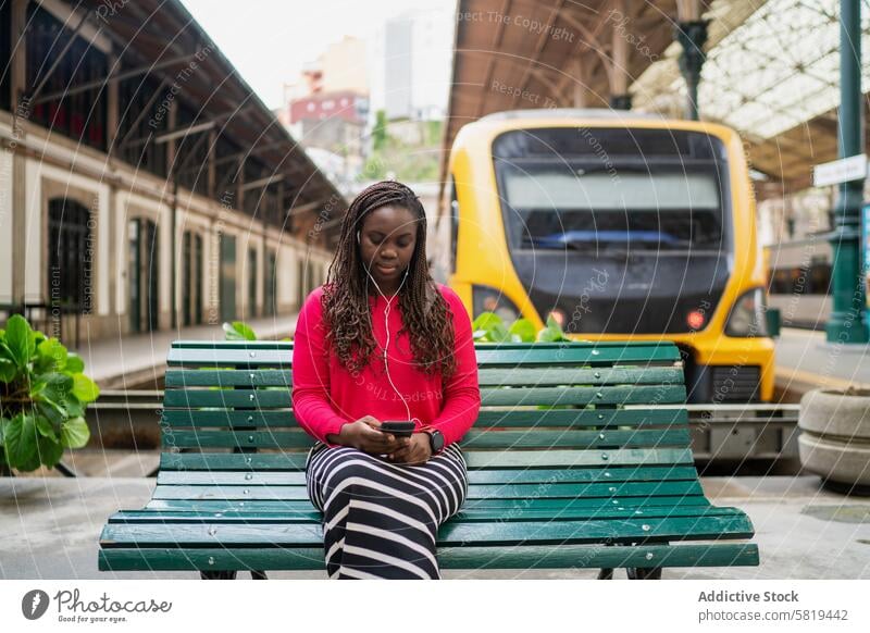 Young black woman using her phone at a train station bench smartphone yellow train sitting green bench engrossed young urban commuter waiting travel