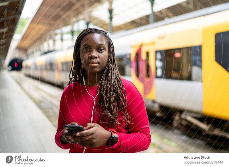 Black woman waiting at train station using smartphone platform travel commuter public transport urban city mobile device listening music red top background