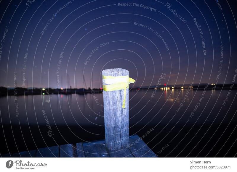 Wooden pole on a jetty leading into a lake at night, lights and a harbor can be seen on the opposite shore Wooden stake Footbridge Body of water Lake Harbour