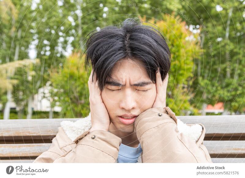 Young man experiencing headache symptoms outdoors discomfort hypoglycemia young asian park bench clutching pain health medical condition suffering