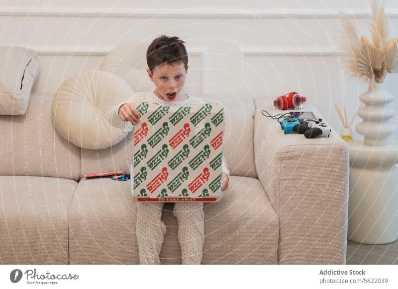 Boy surprised with pizza box at home boy looking at camera excited child couch living room indoor expression pajamas wonder anticipation delivery food snack