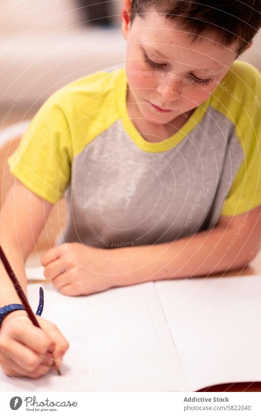 Boy focused on drawing at home boy kid child paper pencil concentrating creative activity indoor looking down eyes closed yellow shirt grey seated hobby art