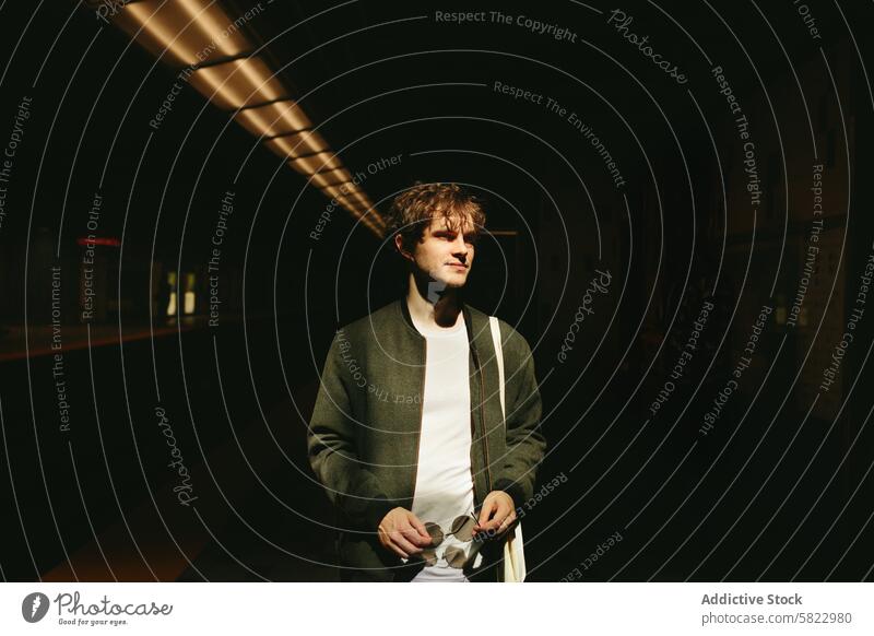 Young man waiting alone at a dimly-lit subway station light shadow casual coat t-shirt sunglasses solitude underground transport urban city commuter travel