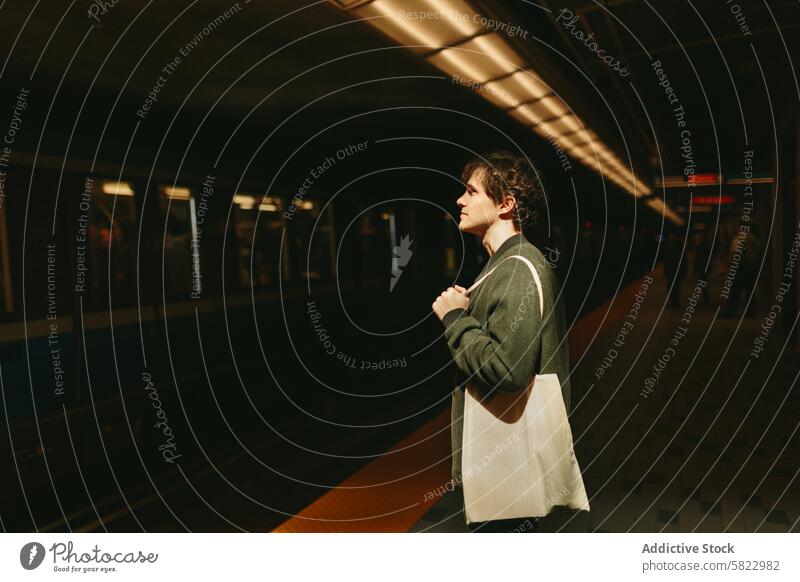 Young man waiting at subway platform during dusk train young male casual jacket tote bag standing thoughtful dim lit sun setting transportation commuter travel