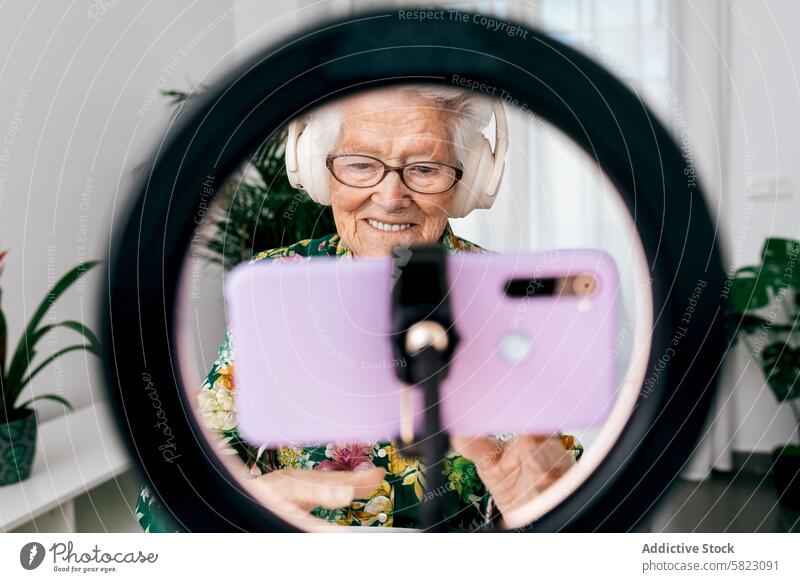 Elderly woman recording a podcast seen through a ring light elderly headphone smartphone influencer home indoor technology digital media content creator video