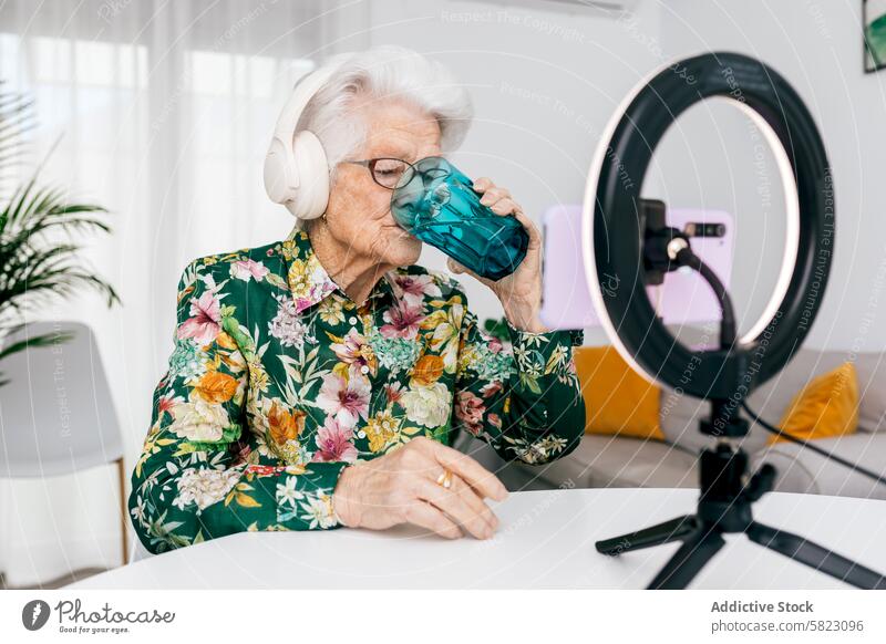 Elderly woman influencer recording a video podcast elderly headphone ring light floral shirt drinking water podcasting technology modern senior media