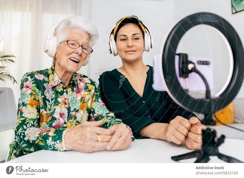 Young and elderly woman recording a podcast together influencer microphone headphones camera desk smiling technology media audio broadcasting talk show