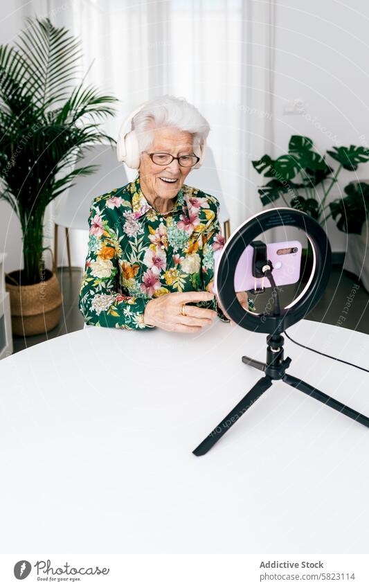 Senior woman records podcast with modern technology senior elderly recording influencer headphones smile joy ring light smartphone mount floral green indoor