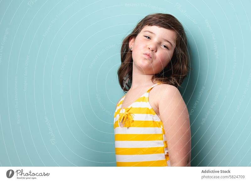 Young girl posing confidently in a striped yellow swimsuit pose playful pout striped swimsuit white pale blue background camera young fashion child expression