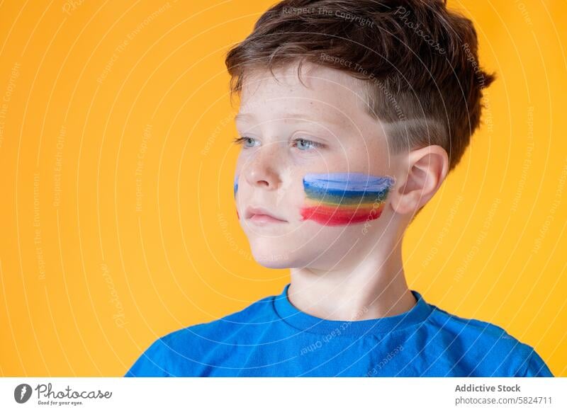 Young boy with colorful flag painted on his cheek child face paint pride contemplation portrait young orange background focused expression culturally diverse