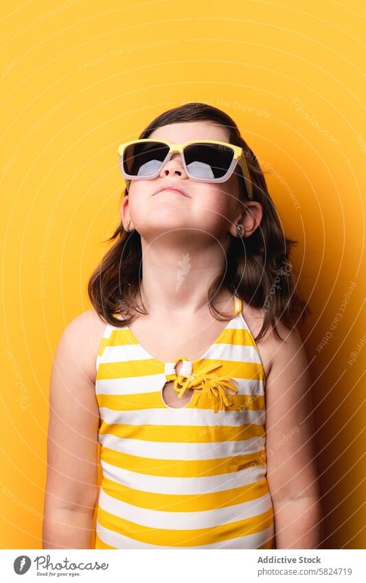 Young girl in sunglasses looking up on yellow background striped swimsuit cheerful stylish upward gaze fashion child young color bright vibrant summer accessory